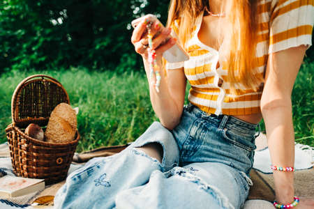 Young woman in stylish summer outfit using technology, sitting on picnic blanket in natural park.の写真素材