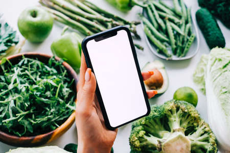 Woman hand holding smartphone with white screen mock-up, in front fresh vegetables on the table. Healthy nutrition menu, or delivery fresh food conceptの写真素材