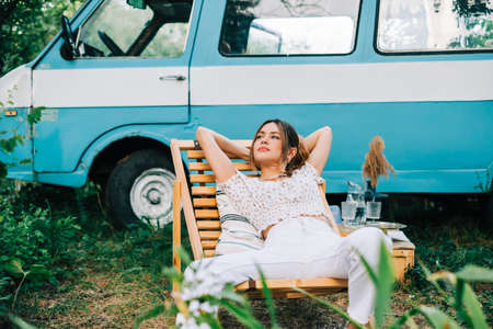 Young woman resting in nature, sitting on wooden chair near van outdoors. Enjoying summer, travel conceptの写真素材