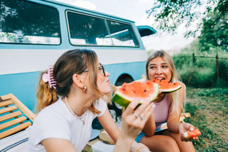 Two young cheerful women friends eating watermelon near van outdoors, summer traveling conceptの写真素材