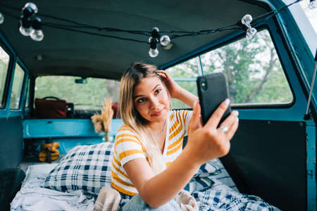 Young woman sitting in a van and using mobile phone, looking on screen, outdoors in nature. Enjoying...