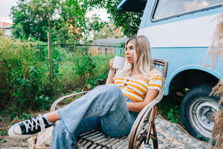 Young woman sitting on wooden chair near van and drinking tea outdoors in nature, during sunset. Enjoying summer, travel conceptの写真素材