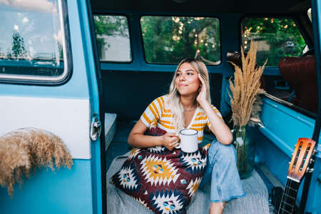 Portrait of happy young woman sitting in a van and drinking tea outdoors in nature, during sunset. Enjoying summer, travel conceptの写真素材