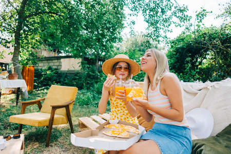 Two women sitting on a sofa in the backyard and eating pizza, drinking lemonade on summer sunny day.