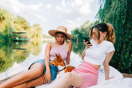 Young woman using mobile phone while resting outdoor in nature near lake with friends and dog.の写真素材