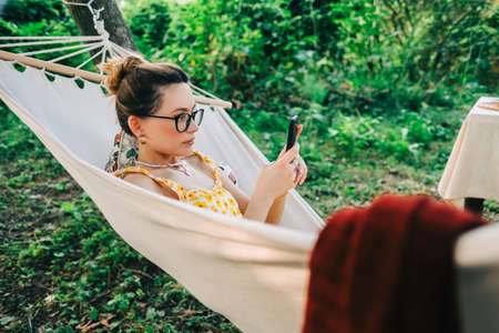 Young woman using mobile phone, relaxing in hammock outdoors, in the backyard garden.の写真素材