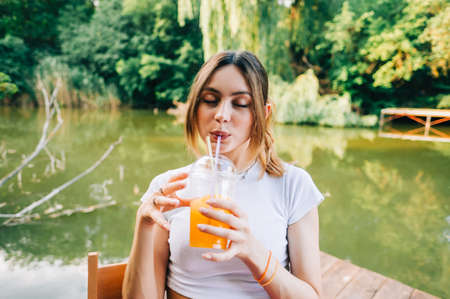 Young attractive woman drinking orange lemonade, resting on a pier on lake outdoor.の写真素材