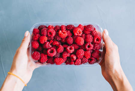 Fresh ripe raspberries in plastic box in hands on blue background, close-up.の写真素材