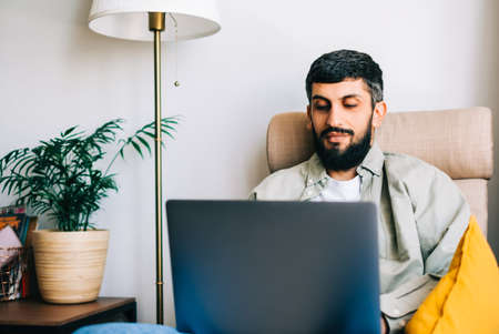 Caucasian man freelancer working at home, sitting on armchair and using laptop computer.