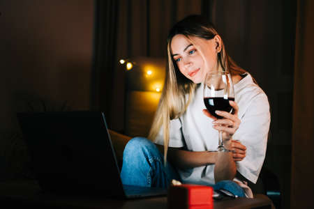Young millennial woman having video call on laptop computer and drinking wine, use technology for communicate with friends or family.の写真素材