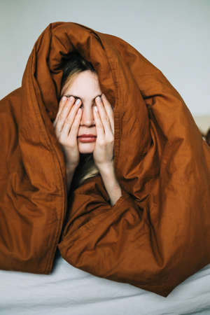 Young caucasian woman waking up in the morning, lying under cover and rubbing face.の写真素材