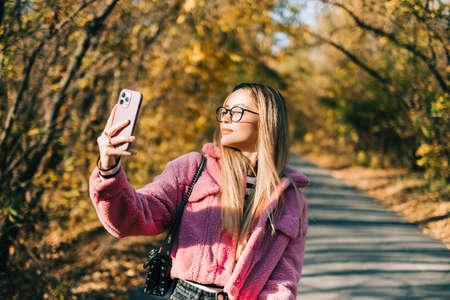 Young woman making self photo and enjoying autumn weather in the park. Woman walking in the autumn park.の写真素材