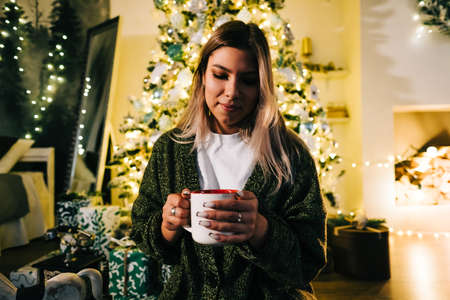 Young caucasian woman relaxing near Christmas tree at home, holding mug with hot drink in Christmas...