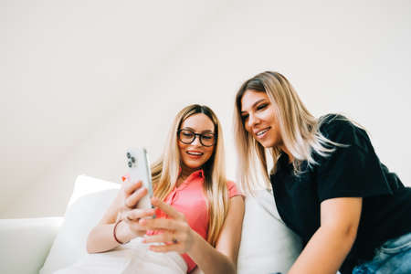Friends heaving fun, sitting on sofa and looking on smartphone screen, two cheerful women together browsing internet.の写真素材