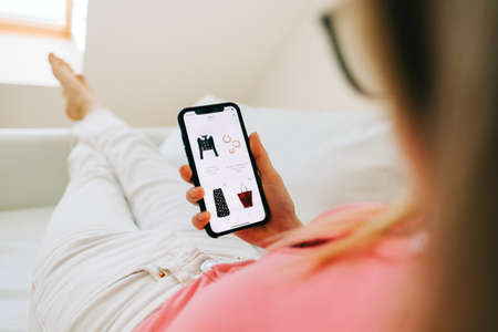 Young woman using online store app buying clothing. Woman at home lying on bed in living room using...
