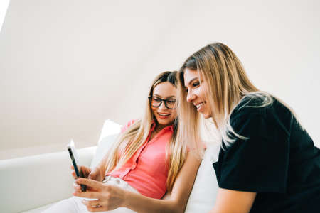 Friends heaving fun, sitting on sofa and looking on smartphone screen, two cheerful women together...
