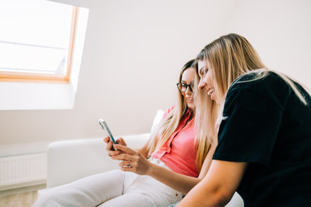 Friends heaving fun, sitting on sofa and looking on smartphone screen, two cheerful women together browsing internet.の写真素材