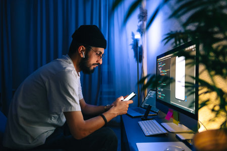 Young man mobile developer writes program code on a computer and check app in smartphone, programmer work in home office.の写真素材