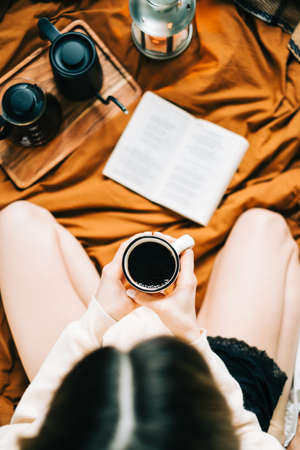 Caucasian woman drinking coffee in the morning and reading book on the bed at home, coziness and comfort concept.の写真素材