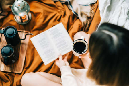 Caucasian woman drinking coffee in the morning and reading book on the bed at home, coziness and...
