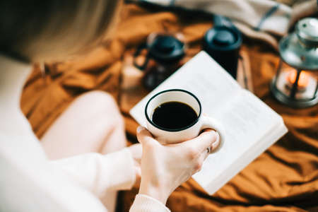 Caucasian woman drinking coffee in the morning and reading book on the bed at home, coziness and comfort concept.の写真素材