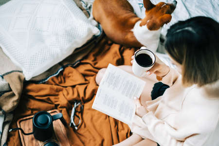 Caucasian woman drinking coffee in the morning and reading book on the bed at home with her pet, coziness and comfort concept.の写真素材