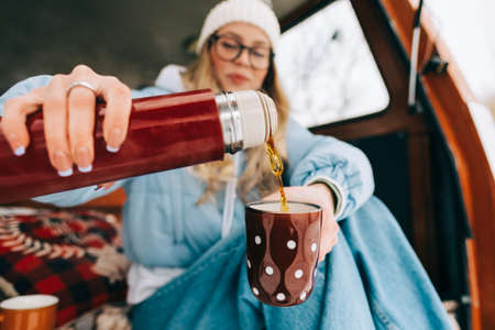 Young caucasian woman pouring hot tea in a cup, sitting in a van in winter camp, enjoying holiday.の写真素材