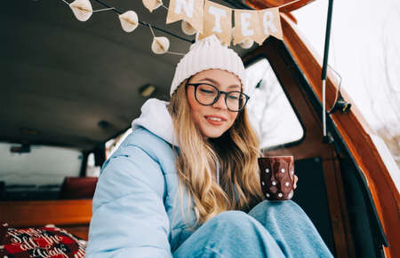 Young caucasian woman drinking tea, sitting in a van in winter camp, enjoying holiday.の写真素材