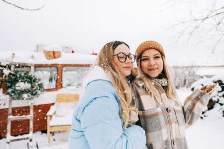 Portrait of two cheerful women friends standing outdoor near van during the snowfall in winter camp.の写真素材