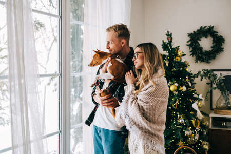 Young couple standing near big window with his dog, enjoying winter holidays.の写真素材