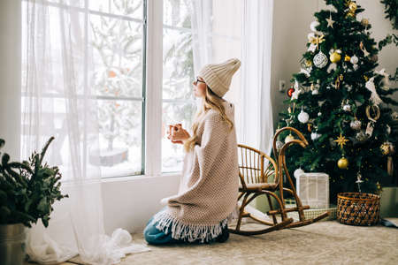 Young caucasian woman standing near window and looking outside in the morning, drinking tea and enjoying winter time.の写真素材