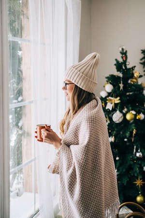 Young caucasian woman standing near window and looking outside in the morning, drinking tea and enjoying winter time.の写真素材