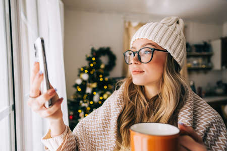 Young caucasian woman standing near window at home and taking photo after snowfall in the morning.の写真素材