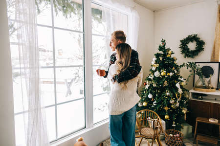 Young couple standing near big window and looking outside, enjoying winter holidays.の写真素材