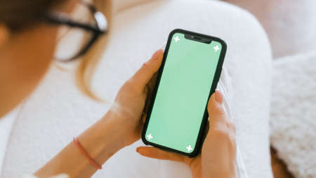 Young woman sitting on sofa at home and using smartphone with green screen.