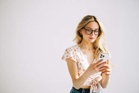 Happy smiling young attractive caucasian woman in eyeglasses using mobile phone on white background with copy space.の写真素材