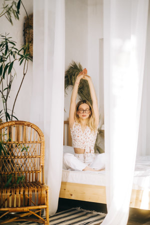 Attractive caucasian young woman in eyeglasses meditate sitting on a bed and enjoying sunny morning.の写真素材