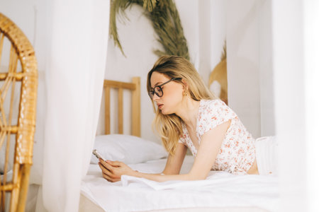 Happy smiling young attractive caucasian woman in eyeglasses using smartphone, lying on a bed.の写真素材