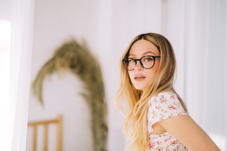 Portrait of attractive caucasian young woman in eyeglasses standing in a bright bedroom in resort.