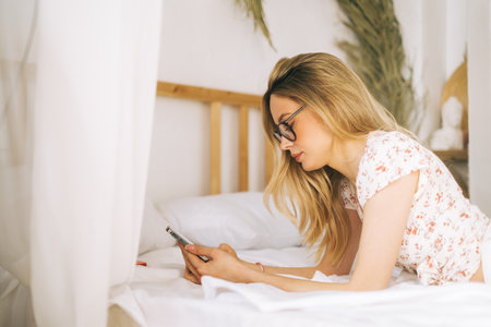 Happy smiling young attractive caucasian woman in eyeglasses using smartphone, lying on a bed.の写真素材