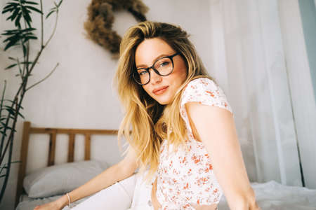 Portrait of attractive young happy caucasian woman in glasses, sitting in cozy bedroom.の写真素材