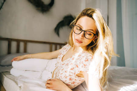 Portrait of attractive young happy caucasian woman in glasses, sitting in cozy bedroom.の写真素材