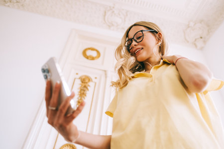 Young caucasian cheerful woman using mobile phone in bright living room, connecting to friends in video call or taking selfie photo.の写真素材