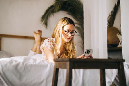 Happy smiling young attractive caucasian woman in eyeglasses using smartphone, sitting on a bed.の写真素材