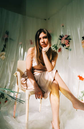 Portrait of attractive caucasian woman in silky dress, sitting in a sunny room with flowers on a wall.の写真素材