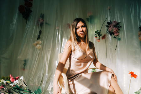 Portrait of attractive caucasian woman in silky dress, sitting in a sunny room with flowers on a wall.の写真素材