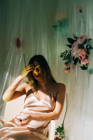 Portrait of attractive caucasian woman in silky dress, sitting in a sunny room with flowers on a wall.の写真素材