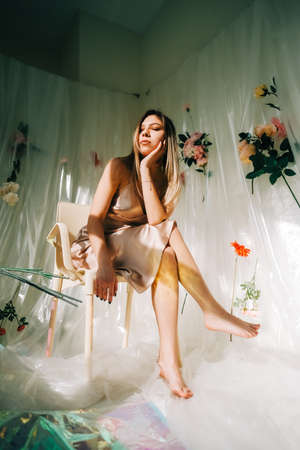 Portrait of attractive caucasian woman in silky dress, sitting in a sunny room with flowers on a wall.の写真素材