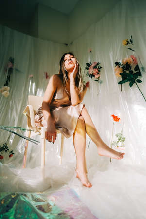 Portrait of attractive caucasian woman in silky dress, sitting in a sunny room with flowers on a wall.の写真素材