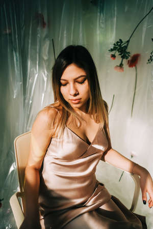 Portrait of attractive caucasian woman in silky dress, sitting in a sunny room with flowers on a wall.の写真素材